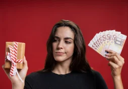 Mujer comparando una caja de regalo y dinero en efectivo, simbolizando la decisión entre gastar o pedir un préstamo a comienzos de año, fondo rojo.