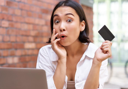 Mulher surpresa segurando cartão de crédito enquanto olha notebook, representando dependência de crédito e preocupação financeira